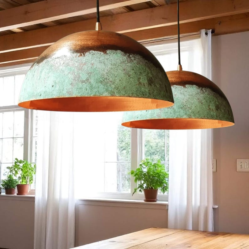 May include: Two dome-shaped pendant lights with a copper-colored interior and a weathered green and brown exterior. The lights hang from black cords with gold-colored accents. The lights are suspended above a wooden table.