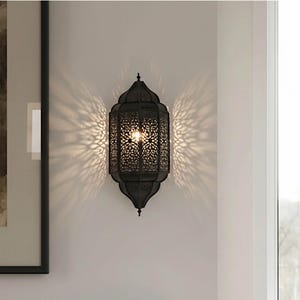 May include: A black metal wall sconce with an intricate, openwork design. The sconce is shaped like a traditional lantern, casting a patterned light on the wall. The light source is visible inside the lantern.