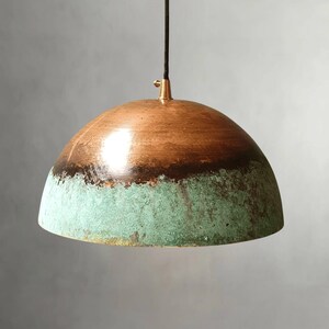 May include: A copper pendant light fixture with a dome-shaped shade. The top half of the shade is a warm copper color, transitioning to a dark brown band, and then to a turquoise-green patina at the bottom. The fixture hangs from a black cord.