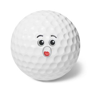 Puede incluir: Una pelota de golf blanca con una cara de dibujos animados sorprendida. La pelota tiene ojos negros, cejas y una boca roja abierta. La superficie con hoyuelos de la pelota es visible.