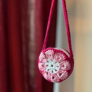 May include: A small, round, crocheted bag in shades of pink and white, with a matching pink strap. The bag features a floral design in the center and is held up by a pink strap. The bag is handmade and has a unique, textured appearance.