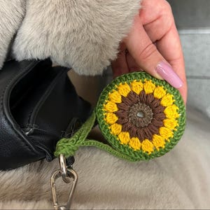 Puede incluir: Un llavero de girasol tejido a ganchillo con colores amarillo, marrón y verde. El llavero redondo tiene un borde verde y una correa verde sujeta a un mosquetón plateado. El llavero lo sostiene una persona.
