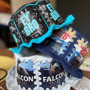 May include: Handmade hats crafted from recycled beer cans. The hats feature crochet brims and stitching, with the can labels visible. One hat displays "EY BRO LJUS LAGER" in blue and white, while another shows "FALCON EXPORT" with a falcon graphic.