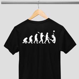 May include: Black t-shirt featuring a white graphic illustrating human evolution, ending with a volleyball player spiking the ball. The design shows a sequence of figures evolving from ape-like forms. The t-shirt is displayed on a wooden hanger.