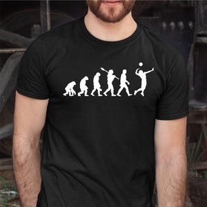 May include: Black t-shirt with a white graphic showing human evolution, ending with a volleyball player. The design progresses from ape-like figures to a volleyball player spiking a ball. The shirt is made of a dark fabric.