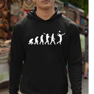 May include: Black hoodie featuring a white graphic illustrating the evolution of volleyball. The design shows human figures transforming into a volleyball player spiking a ball. The hoodie includes a front pocket and a hood.
