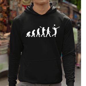 Puede incluir: Sudadera con capucha negra con un gráfico blanco que representa la evolución de un jugador de voleibol. El gráfico muestra una serie de figuras, comenzando con un primate y evolucionando hacia un jugador de voleibol que golpea el balón. La sudadera tiene un bolsillo delantero y una capucha.