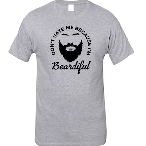 Könnte beinhalten: Hellgraues T-Shirt mit schwarzer Bartgrafik und dem Text "DON'T HATE ME BECAUSE I'M BEARDIFUL" in einem kreisförmigen Design. Das T-Shirt hat einen Rundhalsausschnitt und kurze Ärmel.