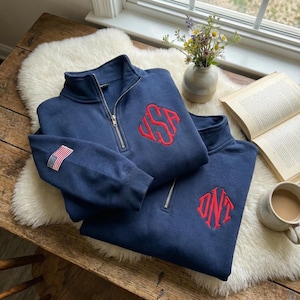 May include: Two navy blue quarter-zip pullovers with red embroidered monograms. One has a small American flag patch on the sleeve. The pullovers are on a white sheepskin rug on a wooden table, with a book, flowers, and a mug.