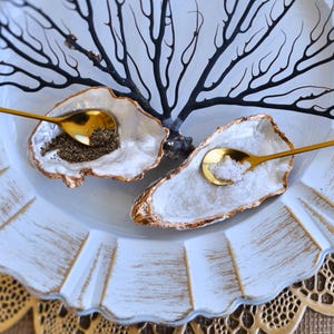 May include: Two gold oyster shell salt cellars with matching gold spoons on a white plate with a black coral branch in the center. The plate is on a gold lace doily.