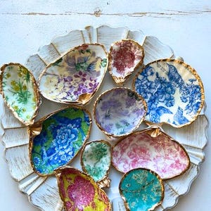 May include: A collection of decorative oyster shell dishes with gold-painted edges, each featuring unique floral and patterned designs in various colors like blue, pink, green, and purple. The shells are arranged on a white, scalloped tray.