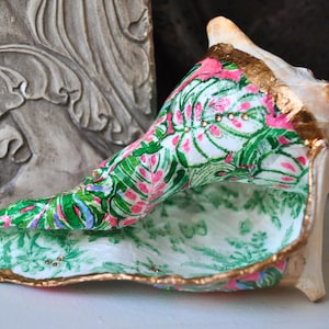 May include: A large, decorated seashell with a gold-painted rim. The interior is lined with a white fabric featuring a green and pink floral pattern. The exterior is a natural cream color.