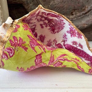 May include: A large seashell with a gold-painted rim, lined with patterned paper. The paper features a lime green and fuchsia floral design on the exterior and a white and fuchsia toile pattern with peacocks and pagodas on the interior.