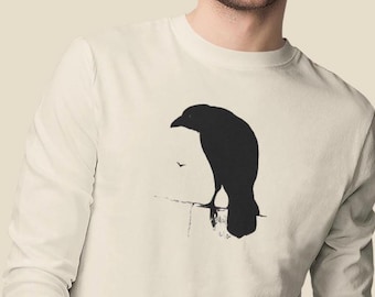 Può includere: Una maglietta bianca a maniche lunghe con una silhouette nera di un corvo sul davanti.