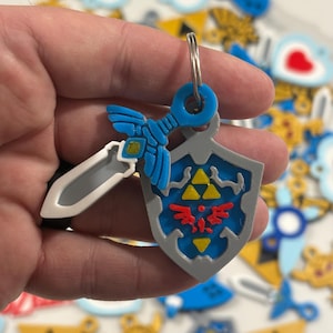 May include: A blue and gray keychain featuring a sword and shield design. The shield has a red emblem and yellow triangles. The keychain is attached to a silver ring. Other similar keychains are in the background.