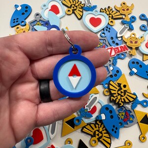 May include: A blue and white keychain with a compass design. The compass has a red needle pointing north. The keychain is surrounded by other keychains with various designs, including hearts, swords, and the Triforce symbol.