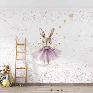 Könnte beinhalten: Eine Aquarell-Illustration eines Kaninchens, das einen lila Tutu trägt und auf einem weißen Hintergrund mit lila und goldenen Akzenten steht.