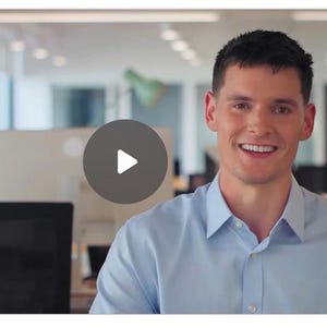 May include: A man in a light blue button-down shirt smiles at the camera. A play button is overlaid on the image. The background is a modern office setting with desks and computer monitors.