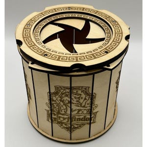 Könnte beinhalten: Eine Holzkiste mit rundem Deckel und sternförmiger Öffnung. Die zylindrische Box zeigt ein Gryffindor-Wappen. Das Design ist lasergeschnitten mit detaillierten Mustern und zeigt eine natürliche Holzoberfläche. Höhe: ca. 15 cm.