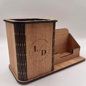 Può includere: Organizzatore da scrivania in legno con portapenne e porta biglietti da visita. L'organizzatore è realizzato in legno marrone chiaro con un design tagliato al laser sui lati. La parte anteriore presenta le lettere "L/D" all'interno di un cerchio. La base è rettangolare.