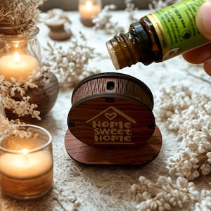 Può includere: Un diffusore di oli essenziali in legno con la scritta "Home Sweet Home". Una piccola bottiglia versa olio nel diffusore. L'immagine include candele accese ed elementi decorativi.