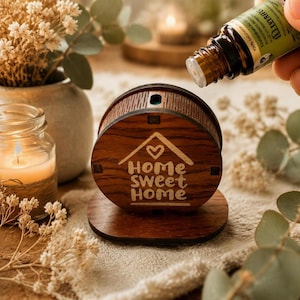 Puede incluir: Un difusor de aceites esenciales circular de madera con acabado marrón oscuro, que presenta un icono de casa grabado, un corazón y el texto "home sweet home" en el frente. Un pequeño frasco de vidrio ámbar se sitúa sobre el difusor.