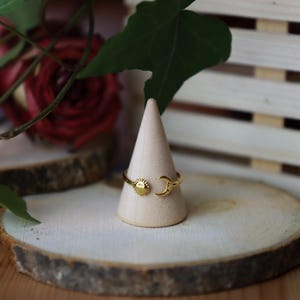 Könnte beinhalten: Ein goldfarbener offener Ring mit einem kleinen Sonnenmotiv an einem Ende und einem Mondsichelmotiv am anderen Ende. Der Ring wird auf einem hellen Holzkegel präsentiert, wobei die himmlischen Designelemente auf dem Band deutlich sichtbar sind.
