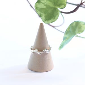 Op de afbeelding: Een zilveren ring met een doorlopend bandontwerp van kleine, gepolijste metalen blaadjes die de vinger omsluiten. De ring wordt getoond op een gladde, beige, kegelvormige standaard, waarbij het gedetailleerde bladmotief zichtbaar is.