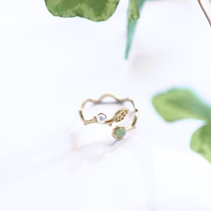 Könnte beinhalten: Ein goldfarbener offener Ring mit einem astförmigen Band, einer kleinen weißen Perle, einem strukturierten Blattdetail und einem runden hellgrünen Stein. Der Ring hat ein filigranes, organisches Design mit polierter Oberfläche und ist verstellbar.