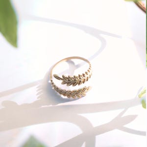 May include: A gold-toned metal ring featuring an open-ended design with two overlapping leaf-shaped branches. The band is thin and circular, with each end of the ring terminating in a detailed, textured leaf motif that wraps around the finger.