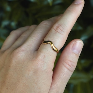 Puede incluir: Un anillo de tono dorado pulido que presenta un diseño liso y continuo en forma de onda que se curva hacia arriba hasta un punto central. La banda tiene un acabado metálico reflectante y un grosor uniforme, rodeando el dedo con fluidez.