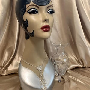 May include: A vintage-style mannequin head with a painted face, dark hair, and red lipstick. It wears a sparkling rhinestone necklace in a tie design and a small earring. A clear glass vase sits beside the mannequin head.