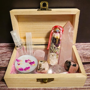 May include: A wooden box with a brass clasp, containing crystals, a small glass bottle with dried flowers, a spray bottle, and a candle with pink rose petals. The box is lined with pink fabric.