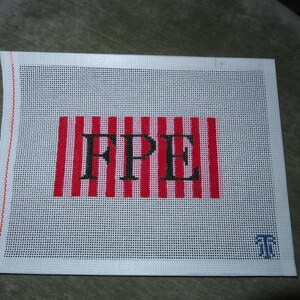 May include: A white needlepoint canvas with a red and white striped rectangle in the center. The letters "FPE" are stitched in black within the rectangle. A small blue emblem is in the lower right corner.