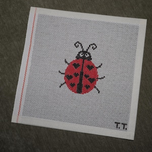 May include: A square, off-white needlepoint canvas featuring a red ladybug with black spots and antennae. The ladybug is centered on the canvas, which has a red stitched border on the left side. The initials "T.T." are in the bottom right corner.