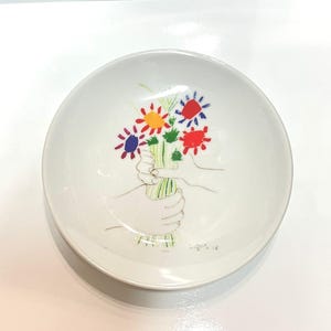May include: White ceramic bowl with a colorful floral design. The bowl features a hand-drawn illustration of a bouquet of flowers in red, yellow, and blue, held by two hands. A unique piece for serving or display.