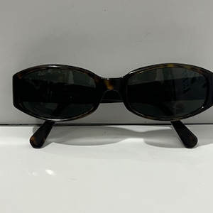 Op de afbeelding: Ovale zonnebril met een montuur van schildpad en donkere glazen. De zonnebril heeft een klassiek design met een licht gebogen bovenkant en een strakke, moderne uitstraling. De pootjes hebben een bijpassend schildpadpatroon.