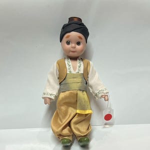 May include: A porcelain doll dressed in a traditional Turkish costume. The doll has brown hair, brown eyes, and a fair complexion. The doll is wearing a white shirt with green embroidery, a gold waistcoat, gold trousers, a yellow sash, and a black turban. The doll is holding a small white tag with a red circle.