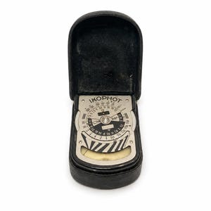 May include: A vintage Ikophot light meter housed in a black leather case. The device features a circular silver dial with black numerical markings, a central rotating scale, and a striped black and white pattern above a small glass window.