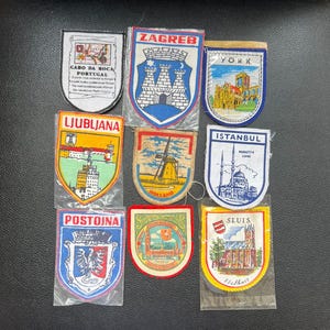 Op de afbeelding: Een verzameling geborduurde reispatches met verschillende stadsnamen en bezienswaardigheden. Patches zijn onder meer "Zagreb", "York", "Ljubljana", "Istanbul", "Postojna" en "Sluis". Elke patch heeft een uniek ontwerp en kleurenschema, met architecturale en culturele elementen.