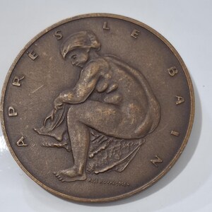 Puede incluir: Una medalla de bronce con una imagen en relieve de una mujer desnuda sentada en una roca. La medalla está inscrita con las palabras "APRES LE BAIN" alrededor del borde.