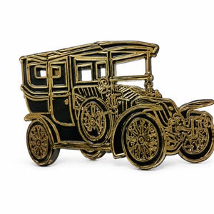 Peut inclure: Une broche en métal en forme d'automobile ancienne, avec un remplissage en émail noir et des contours dorés en relief. Le design représente une voiture classique avec des roues à rayons, une calandre proéminente et une cabine passager carrée.