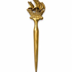May include: A gold-toned metal letter opener featuring a detailed three-masted sailing ship at the top. The handle depicts the ship resting on stylized waves, while the blade tapers to a sharp, pointed tip, creating a functional desk accessory.