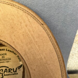 May include: Close-up of a tan leather hat with a stitched brim. The hat has a circular label with text that reads "Best Leather Hats MADE IN AUSTRALIA SINCE 1969". The hat is labeled "CARU Australia" and "SMALL".