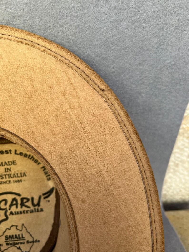 May include: Close-up of a tan leather hat with a stitched brim. The hat has a circular label with text that reads "Best Leather Hats MADE IN AUSTRALIA SINCE 1969". The hat is labeled "CARU Australia" and "SMALL".
