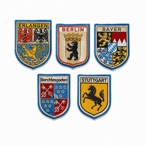 May include: Five shield-shaped embroidered patches featuring German city coats of arms. The patches display the names Erlangen, Berlin, Bayer, Berchtesgaden, and Stuttgart, each with colorful heraldic symbols including eagles, a bear, a horse, and keys.