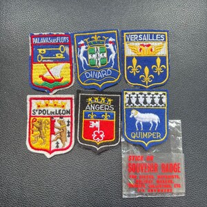 May include: A collection of embroidered souvenir badges with various designs and text. The badges feature city names like Versailles and Quimper, along with images of boats, animals, and heraldic symbols. A clear plastic bag with the text "STICK ON SOUVENIR BADGE" is also present.