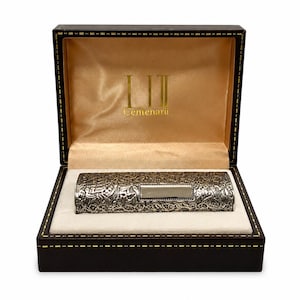 May include: A rectangular, dark brown presentation box with gold stitching, containing a silver-toned, cylindrical object with an intricate, embossed floral pattern and a smooth, rectangular centre plaque. The interior lid features the text "LII Centenarii" in gold.