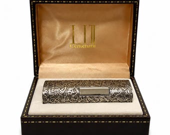 May include: A rectangular, dark brown presentation box with gold stitching, containing a silver-toned, cylindrical object with an intricate, embossed floral pattern and a smooth, rectangular center plaque. The interior lid features the text "LII Centenarii" in gold.