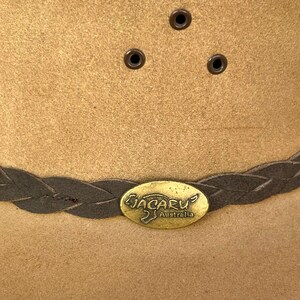 May include: A tan-colored Jacaru Australia hat with a braided band and metal emblem. The hat features three small eyelets on the crown. The hat is made of a suede-like material.